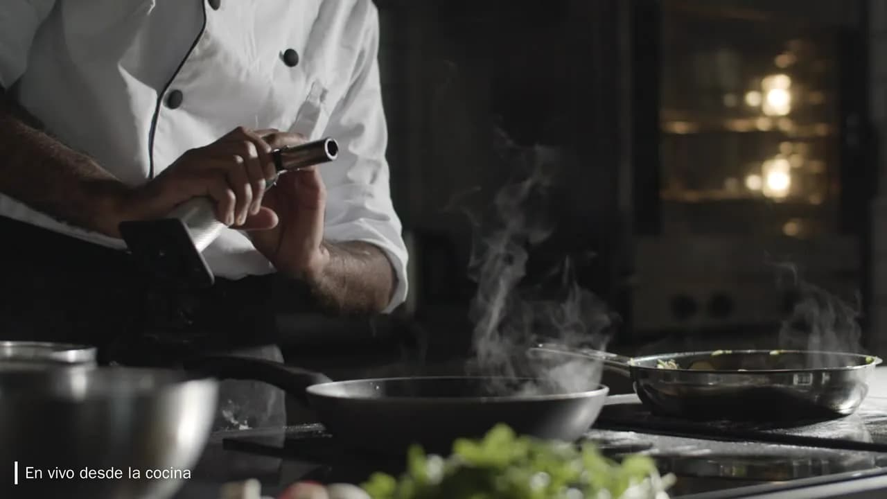 Cámara en vivo: un chef de chaqueta blanca, del pecho para abajo, trabaja junto a sartenes humeantes en la cocina, con el título En vivo desde la cocina